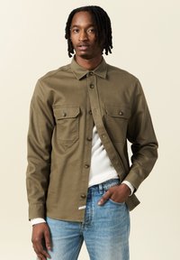 Camicia verde oliva con bottoni, dotata di due tasche sul petto, maniche lunghe e una texture liscia, abbinata a jeans azzurri chiari.