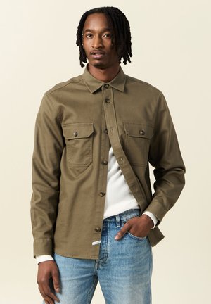 Olivengroene button-up shirt met twee borstzakken, lange mouwen en een soepele textuur, gecombineerd met lichtblauwe jeans.