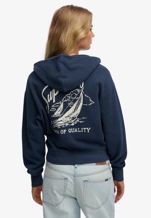 Blonde vrouw die zijwaarts staat, draagt een marineblauwe hoodie met een zeilbootafbeelding en de tekst "Land of Quality", gecombineerd met lichtblauwe spijkerbroek.