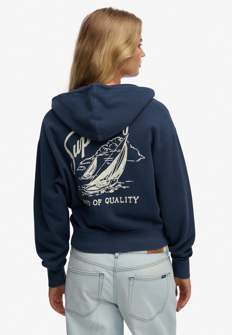 Mujer rubia de perfil que lleva una sudadera con capucha azul marino con un gráfico de velero y el texto "Tierra de Calidad", combinada con jeans azul claro.