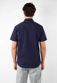 Chemise à manches courtes en tissu bleu marine foncé, présentant un col standard, un empiècement au dos et un branding subtil sur l'épaule. Texture lisse, coupe ajustée.