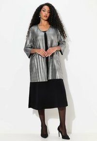 Zilveren geplooide blazer met driekwartmouwen over een zwarte jurk. Hoge hakken maken de look compleet. Soepele textuur en een open voorkant ontwerp.