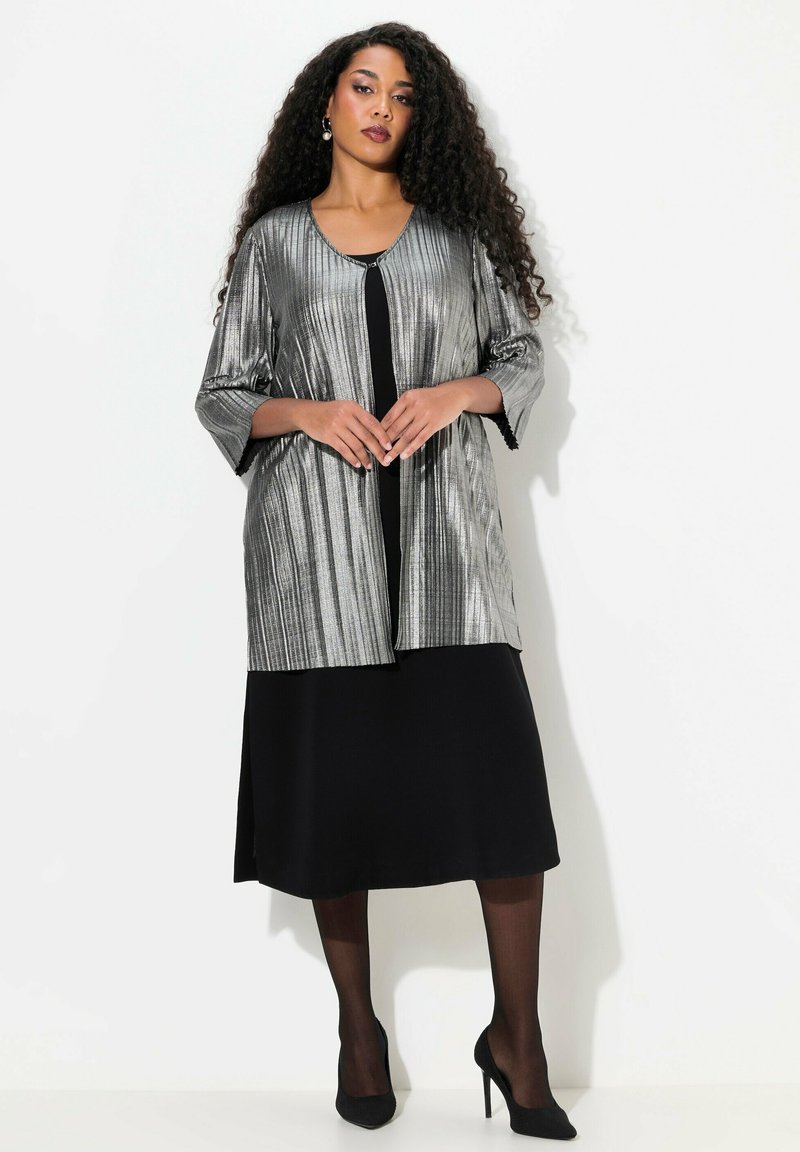 Zilveren geplooide blazer met driekwartmouwen over een zwarte jurk. Hoge hakken maken de look compleet. Soepele textuur en een open voorkant ontwerp.