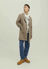 Cappotto in misto lana beige con revers, chiusura a bottoni e tasche laterali, indossato sopra un hoodie bianco, abbinato a jeans blu e stivali marroni.