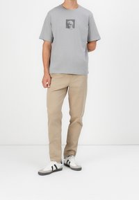 Grå t-shirt med kort ärm med ett dödskallemotiv, tillsammans med beige byxor och vita sneakers med svarta ränder och grå detaljer.