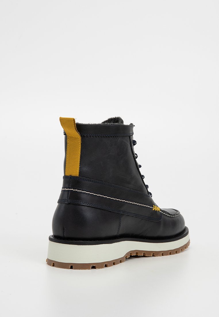 Bottines en cuir noir avec une boucle de couleur jaune, semelle blanche et semelle en caoutchouc brun. Les coutures le long du côté ajoutent de la texture.