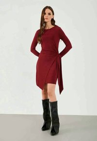 Vrouw in een bordeauxrode jurk met lange mouwen, een asymmetrische zoom en een zij-knoop, gecombineerd met zwarte laarzen tot knielengte op een effen achtergrond.
