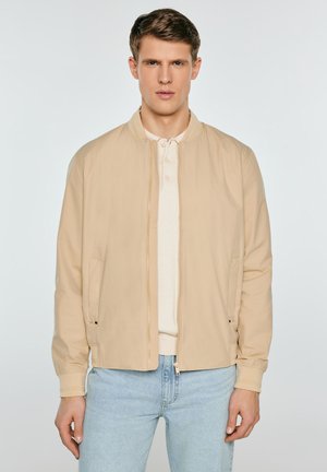 Młody mężczyzna w beżowej kurtce na zamek błyskawiczny, pod którą ma jasną koszulkę polo, oraz w jasnoniebieskich dżinsach, stoi na tle gładkiej białej ściany.