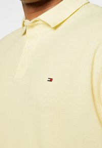 Polo shirt jaune en tissu texturé, avec col et patte de boutonnage à trois boutons. Présente un logo brodé avec des accents rouge, blanc et bleu.