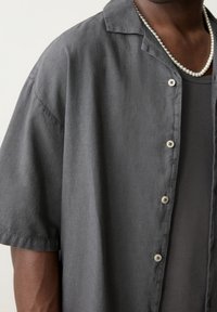 Primer plano de una persona vistiendo una camisa de botones de manga corta en gris oscuro sobre una camiseta sin mangas oscura, adornada con un collar de perlas.