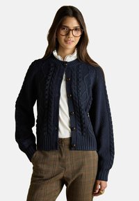 Jūras flotes krāsas adīts džemperis ar strukturāliem kabeļu rakstiem, apaļu apkakli un pogu aizdari; apvienots ar raibām biksēm. Zem tā valkā balto kreklu.