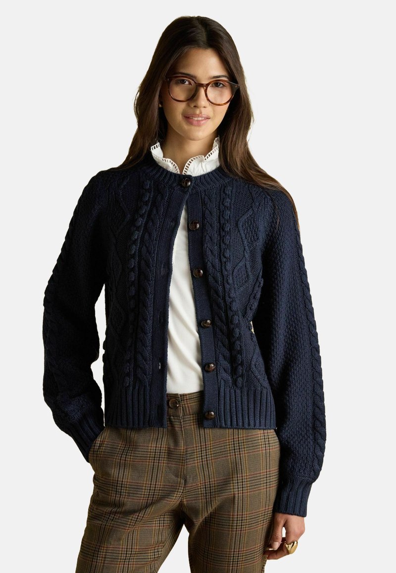 Jūras flotes krāsas adīts džemperis ar strukturāliem kabeļu rakstiem, apaļu apkakli un pogu aizdari; apvienots ar raibām biksēm. Zem tā valkā balto kreklu.