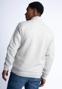 Ljust grått strukturerat sweatshirt med hög krage, ribbad ärmslut och fåll. Bakifrån visar den en slät finish och subtilt mönster.