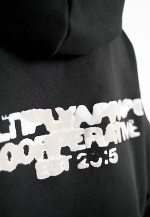 Sudadera negra con capucha, con un texto blanco desgastado que dice: "MULTYARP COOPERATIVE EST 2015", resaltando una tela texturizada.