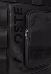Sac à dos noir texturé avec poche avant zippée et le mot embossé "LACOSTE" verticalement sur le panneau latéral.