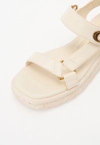 Beige platformová sandál s dvěma popruhy, zlatými kovovými doplňky a embosovanými detaily na klínové podrážce. Hladký povrch.