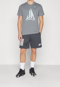 Camiseta deportiva gris con logo reflectante, combinada con pantalones cortos gris oscuro con letras blancas. Zapatillas deportivas negras y calcetines blancos.