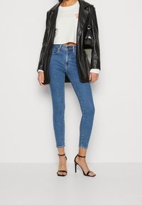 Black faux leather blazer over a cropped white shirt with red text, paired with high-waisted blue skinny jeans and black strappy heels.