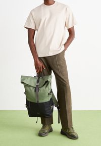 Mies, jolla on päällään vaaleanbeige t-paita, ruskeat pinstripe-housut ja vihreät kengät, pitää kädessään vihreää ja mustaa rullaparista reppua.