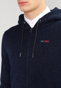 Marineblauwe hoodie met ritssluiting, capuchon met trekkoord, gestructureerde stof en een klein logo in rood en wit op de borst.