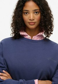 Granatowa bluza z prążkowanym dekoltem i wyhaftowanym logo "Superdry", założona na jasnoróżową koszulę w białe paski z kołnierzykiem.