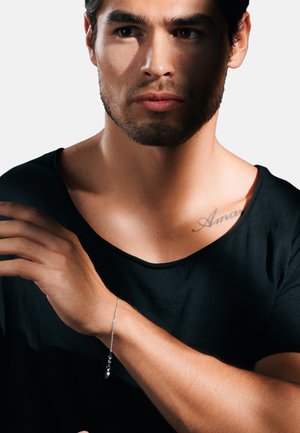 Silbernes Armband mit einem kleinen runden Anhänger, das an einem nackten Handgelenk getragen wird, vor einem dunklen T-Shirt und sanft beleuchteten Gesichtszügen.