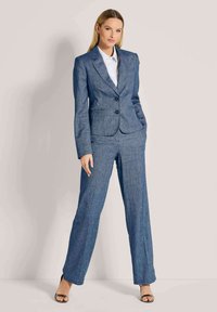 Vrouw staat tegen een effen muur, gekleed in een aansluitend blauwe broekpak met een witte blouse en zwarte open-toe hakken, met de handen ontspannen aan de zijkanten.