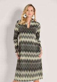 Frau trägt ein langärmliges Midi-Kleid mit Zickzackmuster in Beige, Schwarz und Grau, mit großen weißen abstrakten Ohrringen, vor einem schlichten Hintergrund stehend.
