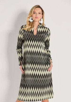 Frau trägt ein langärmliges Midi-Kleid mit Zickzackmuster in Beige, Schwarz und Grau, mit großen weißen abstrakten Ohrringen, vor einem schlichten Hintergrund stehend.
