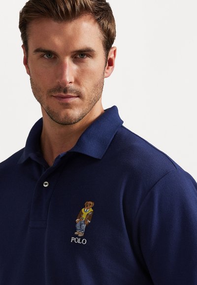 Tumši zila polo krekls, kas izgatavots no teksturētas auduma, ar krāsainu izšūtu lāča logotipu un "POLO" tekstu kreisajā krūtī.