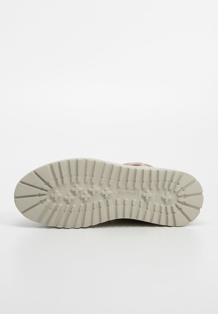 Podeszwa buta w jasnobeżowej gumie z teksturowanym wzorem grip, zawierająca poziome i pionowe rowki oraz wytłoczonym logo na środku.