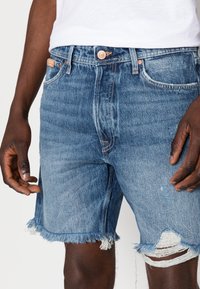 Närbild av en person som bär ljusblå denimshorts med fransade kanter och ett hål på höger ben, kombinerat med en vit skjorta.