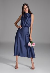 Robe en satin bleu marine avec un col haut et un design sans manches. Présente une taille froncée et descend jusqu'au milieu du mollet. Le mannequin tient des fleurs roses.
