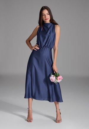 Marineblaues Satin-Kleid mit hohem Kragen und ärmellosem Design. Verfügt über eine geraffte Taille und fließt bis zur Mitte der Waden. Das Model hält pinkfarbene Blumen.
