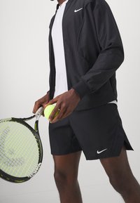 Černá sportovní bunda a šortky Nike, bílá košile s límečkem, držící žlutý tenisový míček a černou tenisovou raketu se zelenými akcenty.