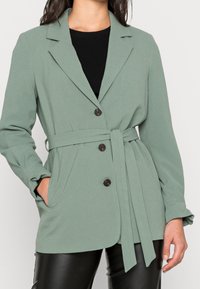 Cappotto verde con cintura, in tessuto testurizzato, colletto con risvolto, tre bottoni marroni e due tasche laterali, abbinato a una maglietta e pantaloni neri.