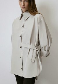 Camicia button-up grigio chiaro con colletto a punta, maniche a tre quarti e dettaglio in vita legato. Presenta bottoni scuri e una texture di tessuto liscia.