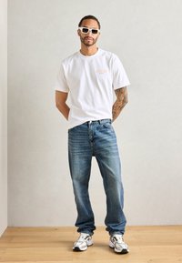 Witte katoenen t-shirt met klein oranje logo, blauwe denim jeans en grijze sneakers met zwarte accenten. Vierkante witte zonnebril maakt de look compleet.