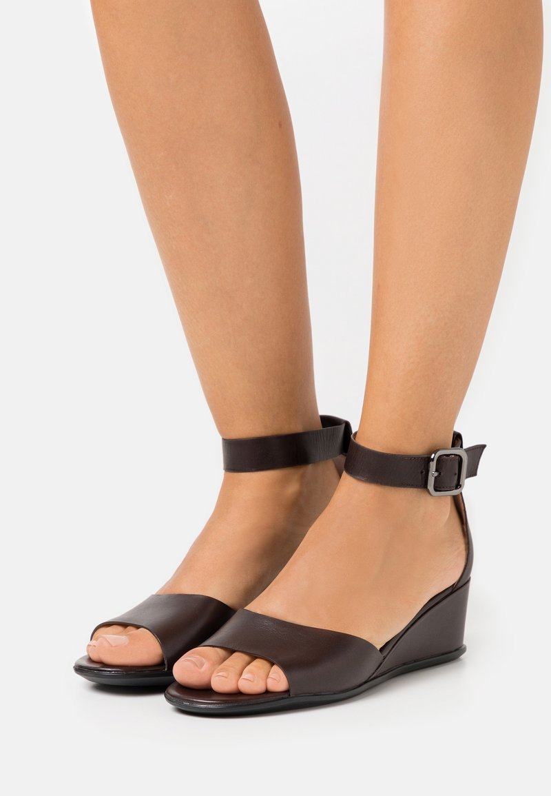 Pieds chaussés de sandales compensées en cuir marron foncé à bouts ouverts et brides à la cheville avec boucles argentées sur fond blanc.