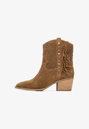 Bota de tobillo de ante marrón con diseño floral recortado, detalle de flecos y tachuelas metálicas. Cuenta con tacón apilado y punta puntiaguda.