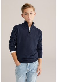 Szőke fiú kék szemekkel, sötétkék negyed cipzáras pulóvert és világoskék farmert viselve, egyik kezét a zsebében tartva áll.