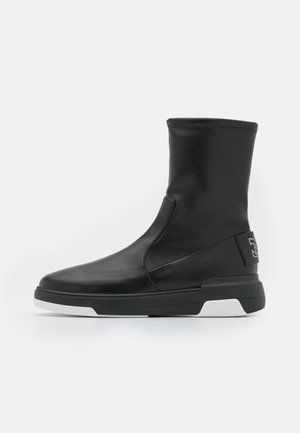 Botte noire mi-mollet en cuir lisse, avec bout arrondi, semelle plate noire avec accents blancs, et languette cousue à l'arrière du talon.