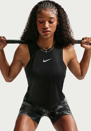 Жена в черен Nike потник и камуфлажни къси панталони държи щанга на раменете си в клекнала позиция на едноцветен светъл фон.