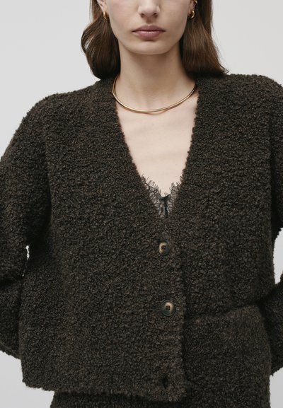 Cardigan en maille brun texturé avec un col en V, trois boutons et une coupe courte, associé à un haut avec dentelle en dessous.
