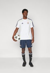 Camiseta blanca de Adidas con acentos amarillos y pantalones cortos azul marino, calcetas negras con rayas blancas, botas negras, sosteniendo un balón de fútbol con un diseño.