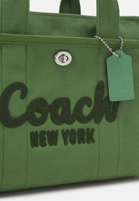 Grön canvasväska med präglad "Coach"-logotyp, silverdetaljer och en grön etikett med texten "COACH NEW YORK." Texturerat tyg.