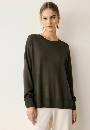 Frau trägt einen dunkelgrünen Pullover mit langen Ärmeln, weiße Hose und große goldene Ohrringe, steht vor einem schlichten hellen Hintergrund.