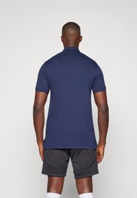 Marineblauwe polo met een kraag, korte mouwen en een rechte zoom. Gladde textuur, gedragen met donkere shorts. Minimalistisch ontwerp.