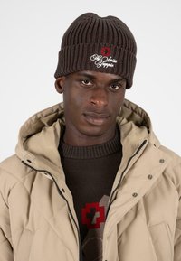 Bruine gebreide beanie met ribbeltextuur en geborduurde rode kruis en witte tekst. Draagt hij met een beige gewatteerde jas en een bruine trui.