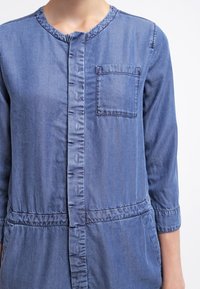 Ljusblå denimskjorta med rund halsringning, knappstängning framtill, en bröstficka och gathered midjedetalj. Långa ärmar.